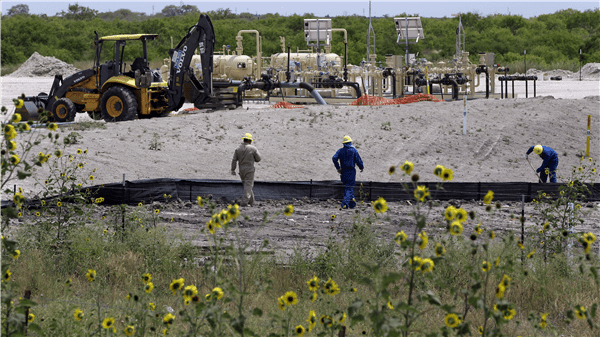 Two men in blue coveralls and yellow hats standing on a dirt road.